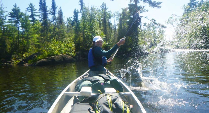 From the back of a canoe, the person in front turns around to splash water at the person taking the photo.